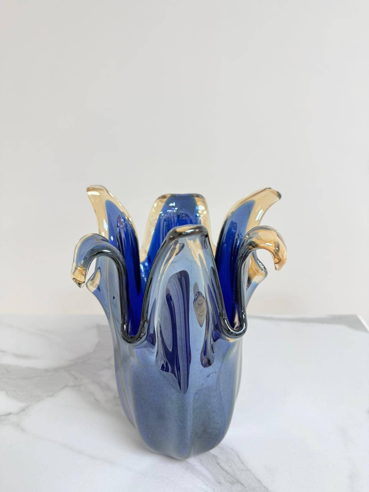 Der Murano-Traum | Die mittelgroße „Giusy“-Vase aus blauem Murano-Glas (H 24, Ø 19 cm) mit wellenförmigem, blütenähnlichem Rand in Blau und Bernstein präsentiert edle italienische Glaskunst auf weißem Marmor vor hellem Hintergrund.