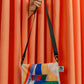 Eine Hand, die aus einem orangefarbenen Vorhang auftaucht, hält die Notabag 'Flow' Leichte Umhängetasche - eine farbenfrohe, rechteckige Umhängetasche von Notabag mit verstellbarem schwarzen Riemen und einem geometrischen Muster in Blau, Rot, Gelb, Grün und Weiß.