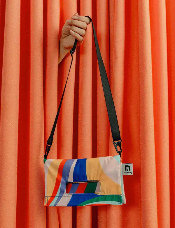 Eine Hand, die aus einem orangefarbenen Vorhang auftaucht, hält die Notabag 'Flow' Leichte Umhängetasche - eine farbenfrohe, rechteckige Umhängetasche von Notabag mit verstellbarem schwarzen Riemen und einem geometrischen Muster in Blau, Rot, Gelb, Grün und Weiß.