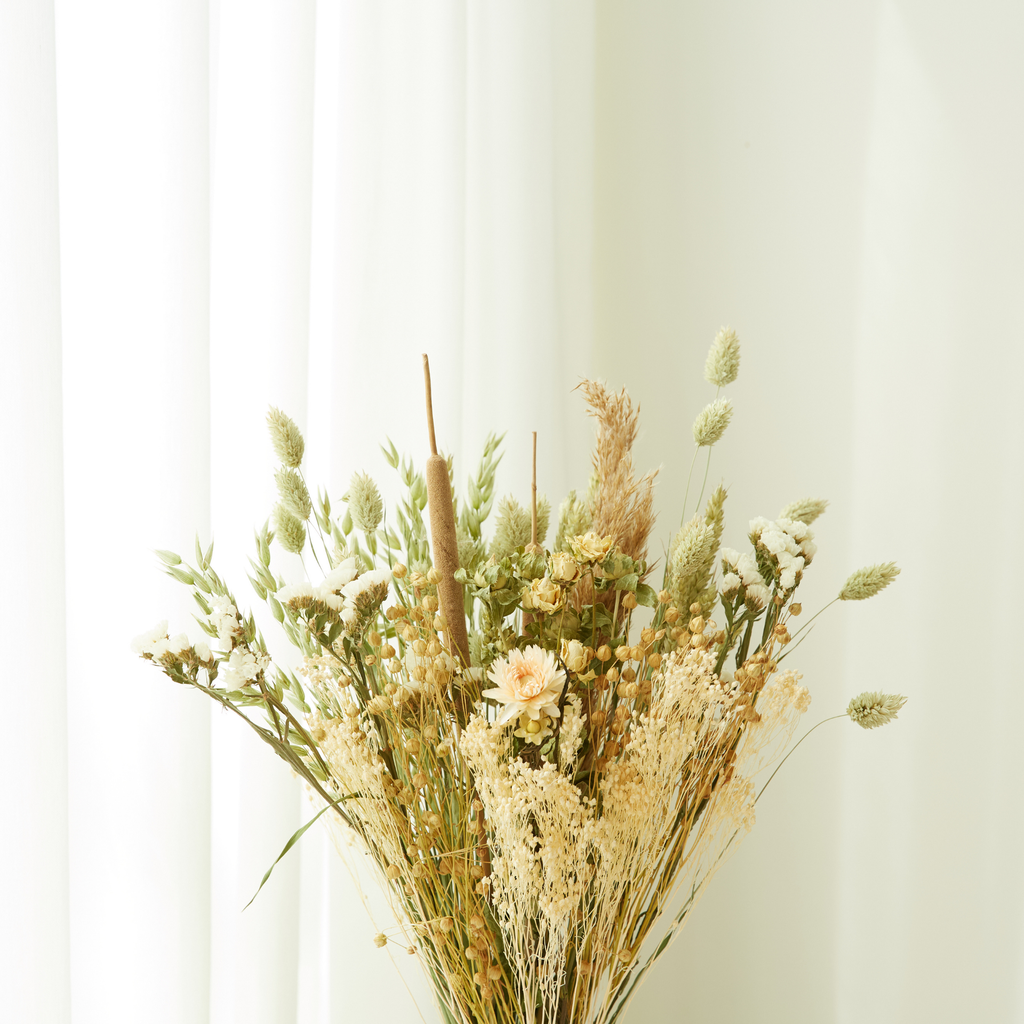 Ein Strauß aus getrockneten Feldblumen, das Modell „Natur“ in Größe M von Wildflowers by Floriette, in sanften Beige- und Grüntönen, ist vor transparenten weißen Vorhängen arrangiert und schafft so eine ruhige und minimalistische Atmosphäre.