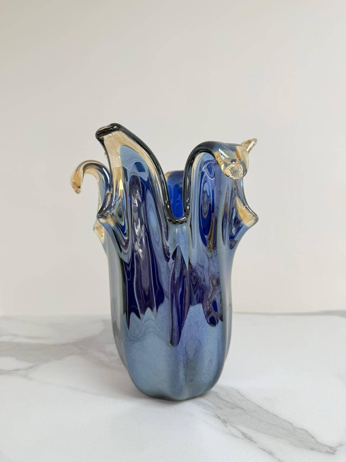 Die Murano Dream – eine mittelgroße „Giusy“-Vase aus blauem Murano-Glas (H 24, Ø 19 cm) mit gewellten Rändern – steht auf weißem Marmor und präsentiert feine italienische Glaskunst vor einem schlichten, hellen Hintergrund.