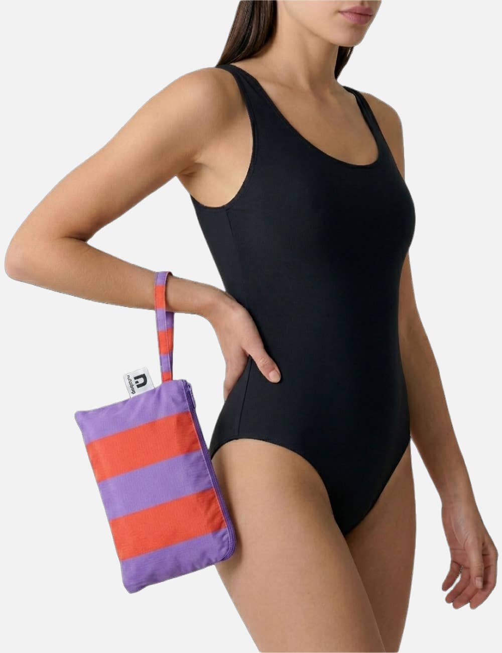 Eine Frau in einem schwarzen, ärmellosen Badeanzug hält eine Notabag | Handtuch rot & lila gestreift 78 × 160 cm am kurzen Riemen an ihrem Oberschenkel fest; das Bild, das oberhalb ihrer Knie und unterhalb ihrer Augen beschnitten ist, zeigt die gestreifte Handtuchtasche von Notabag.
