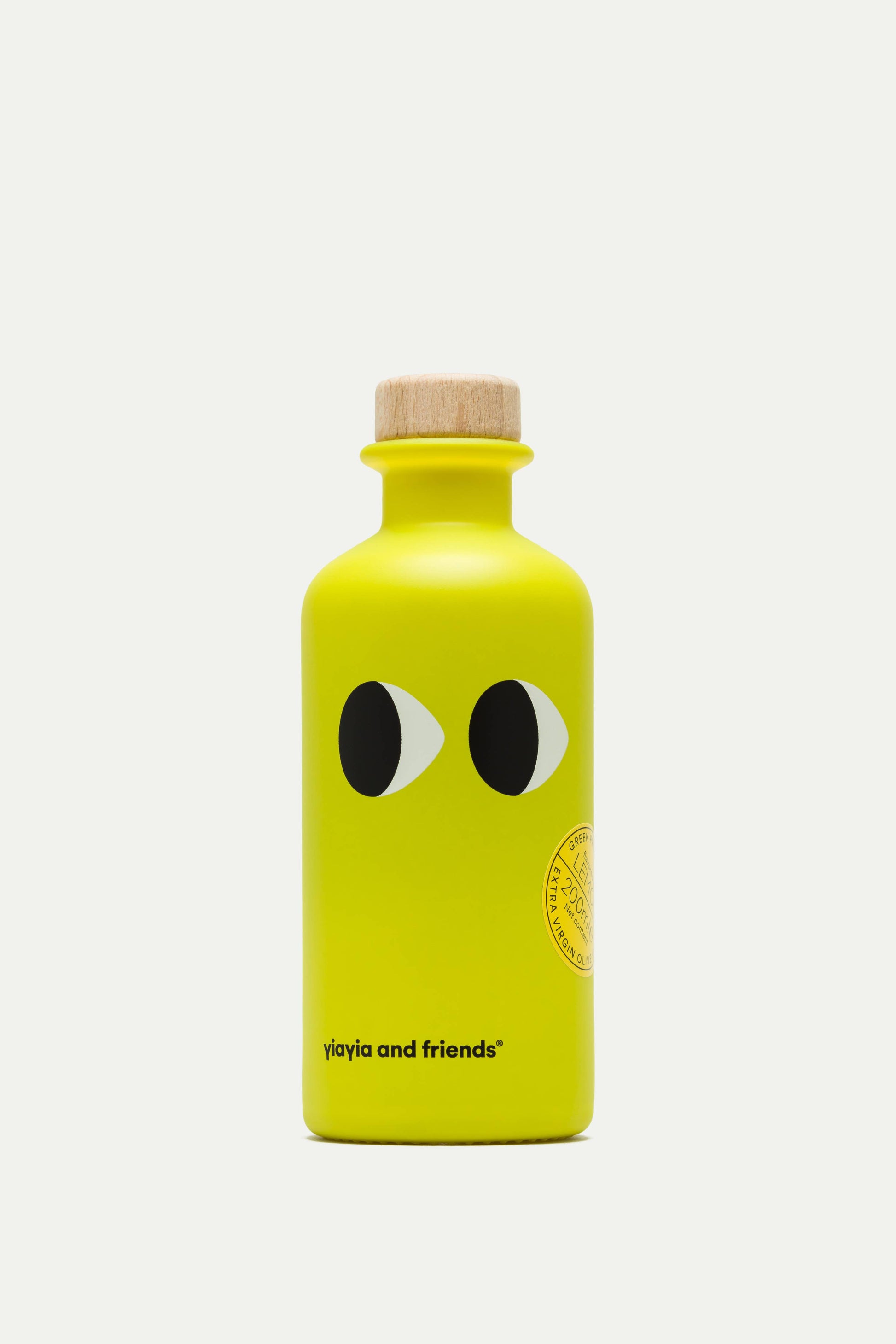 Die leuchtend gelbe 200-ml-Flasche von yiayia and friends Natives Olivenöl extra mit Zitrone besticht durch verspielte, cartoonartige Augen, einen Holzverschluss und ein minimalistisches Design mit dem Schriftzug "yiayia and friends" am Boden - ein Hinweis auf frische Zitronennoten.