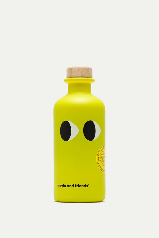 Die leuchtend gelbe 200-ml-Flasche von yiayia and friends Natives Olivenöl extra mit Zitrone besticht durch verspielte, cartoonartige Augen, einen Holzverschluss und ein minimalistisches Design mit dem Schriftzug "yiayia and friends" am Boden - ein Hinweis auf frische Zitronennoten.