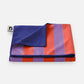 Das „Notabag | Handtuch rot & lila gestreift 78 × 160 cm“ von Notabag ist ein gefaltetes Handtuch mit breiten roten und violetten Streifen, dunkelblauer Rückseite, aus recyceltem Material, schnell trocknend und mit einem Logo-Etikett, abgebildet auf einem schlichten weißen Hintergrund.