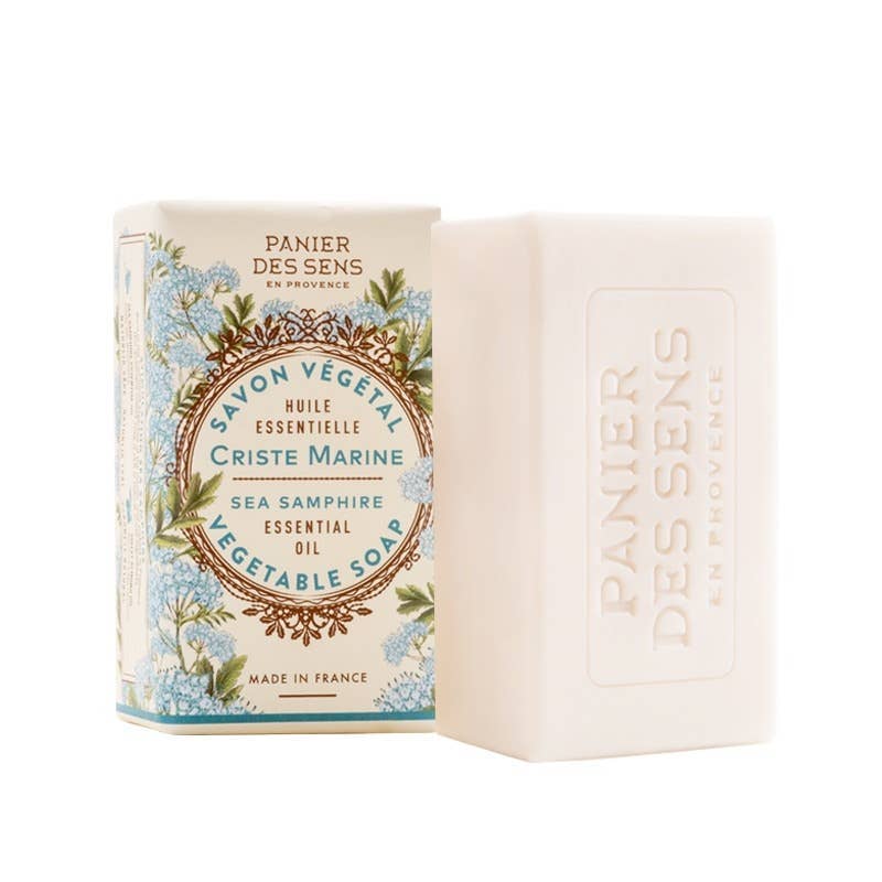 Ein Stück Panier Des Sens | Pflanzliche Seife Meerfenchel 150 g steht neben seiner dekorativen Schachtel mit floralen Motiven und sanften blauen Akzenten. Sowohl auf der Seife als auch auf der Verpackung steht der Markenname Panier Des Sens auf Französisch und Englisch.