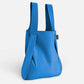 Eine Notabag | „Original Blue“-Tasche, Rucksack und Handtasche in einem von Notabag steht aufrecht vor einem hellen Hintergrund. Die moderne, wiederverwendbare Tasche hat ein schlichtes Design und ein kleines schwarzes Etikett an der Seite.