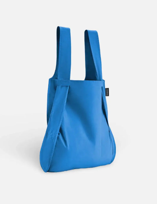Eine Notabag | „Original Blue“-Tasche, Rucksack und Handtasche in einem von Notabag steht aufrecht vor einem hellen Hintergrund. Die moderne, wiederverwendbare Tasche hat ein schlichtes Design und ein kleines schwarzes Etikett an der Seite.