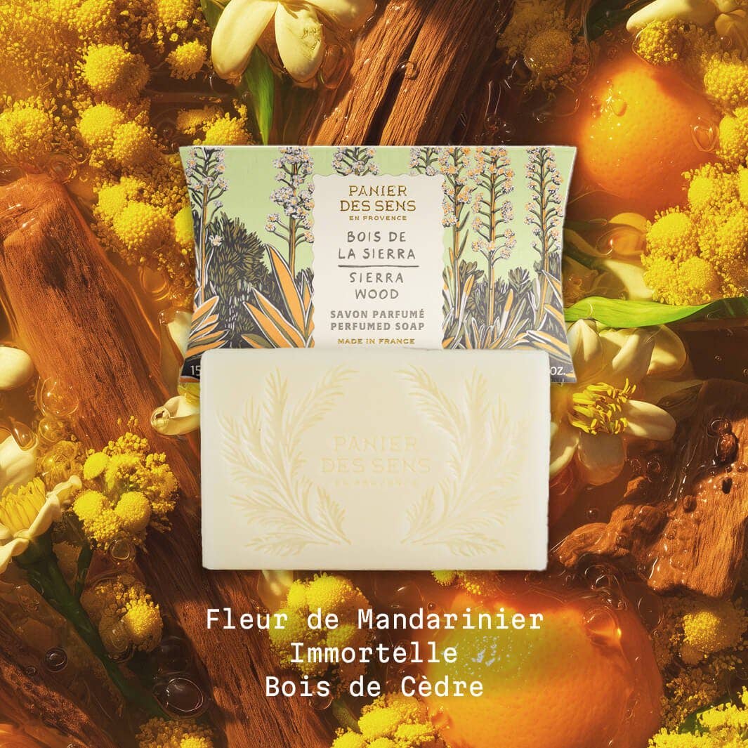 Ein Stück Panier Des Sens Duftseife Sierraholz 150 g ruht in seiner dekorativen Schachtel, umgeben von Orangenscheiben, gelben Blüten und Holz. Diese französische Pflanzenseife hat einen holzig-würzigen Duft mit Mandarinenblüten und Zedernholz.