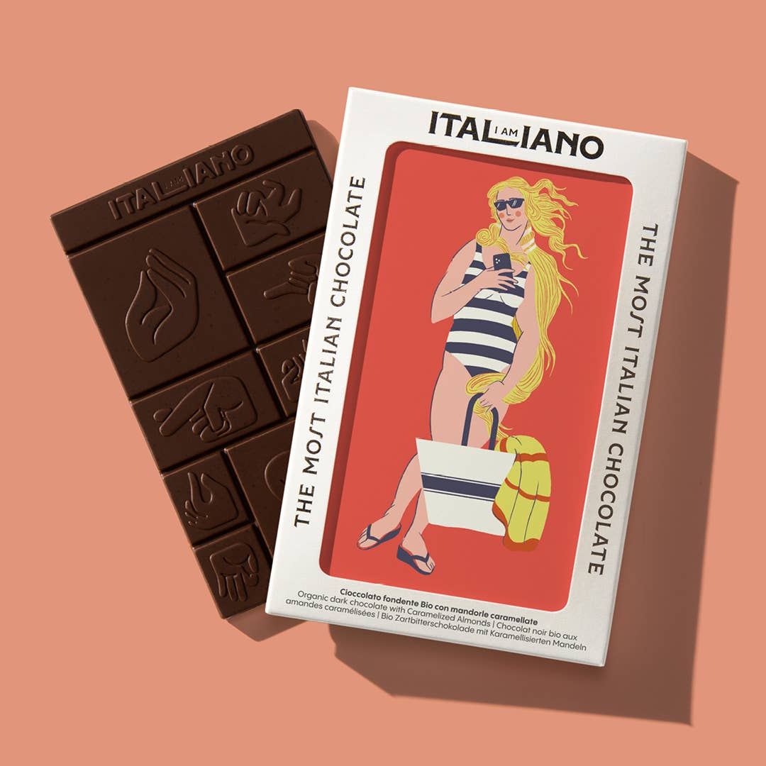 Ein Riegel I AM ITALIANO | 'Venere' Bio Zartbitterschokolade mit Mandel neben seiner Verpackung, die eine blonde Frau im Badeanzug mit Sonnenbrille zeigt, die eine Tasche und ein gelbes Handtuch hält. Auf der Verpackung steht ITALIANO.