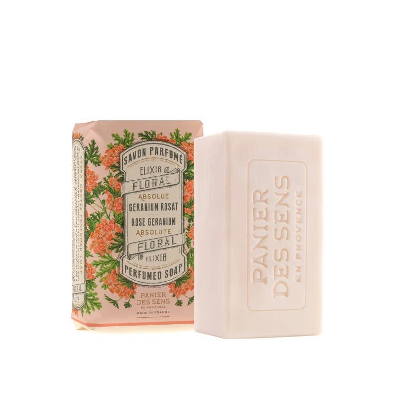 Ein Stück Panier Des Sens | Duftseife Rosengeranie 150 g liegt neben seiner dekorativen Schachtel mit orangefarbenen und grünen botanischen Motiven und dem Text "Savon Parfumé Floral Elixir". Die Seife ist mit der Marke Panier Des Sens geprägt.