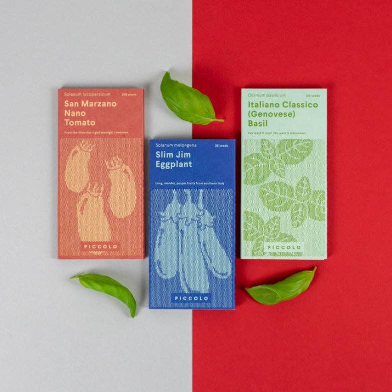 Drei farbenfrohe Saatgutpakete von Piccolo Seeds | Kollektion 'Aubergine Parmigiana' - rot für San Marzano Nano Tomate, blau für Slim Jim Aubergine, grün für Italiano Classico Basilikum - sind auf einer grauen Fläche arrangiert, auf der drei Basilikumblätter verstreut sind.