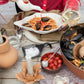 Eine Person in einem roten Hemd gießt Soße auf Spaghetti mit Muscheln, die auf der BYON | Design Schale Alima von Sagaform GmbH serviert werden, zusammen mit Weißwein, Kirschtomaten, Salz, Knoblauch und einer Kerze auf einem Holztisch.