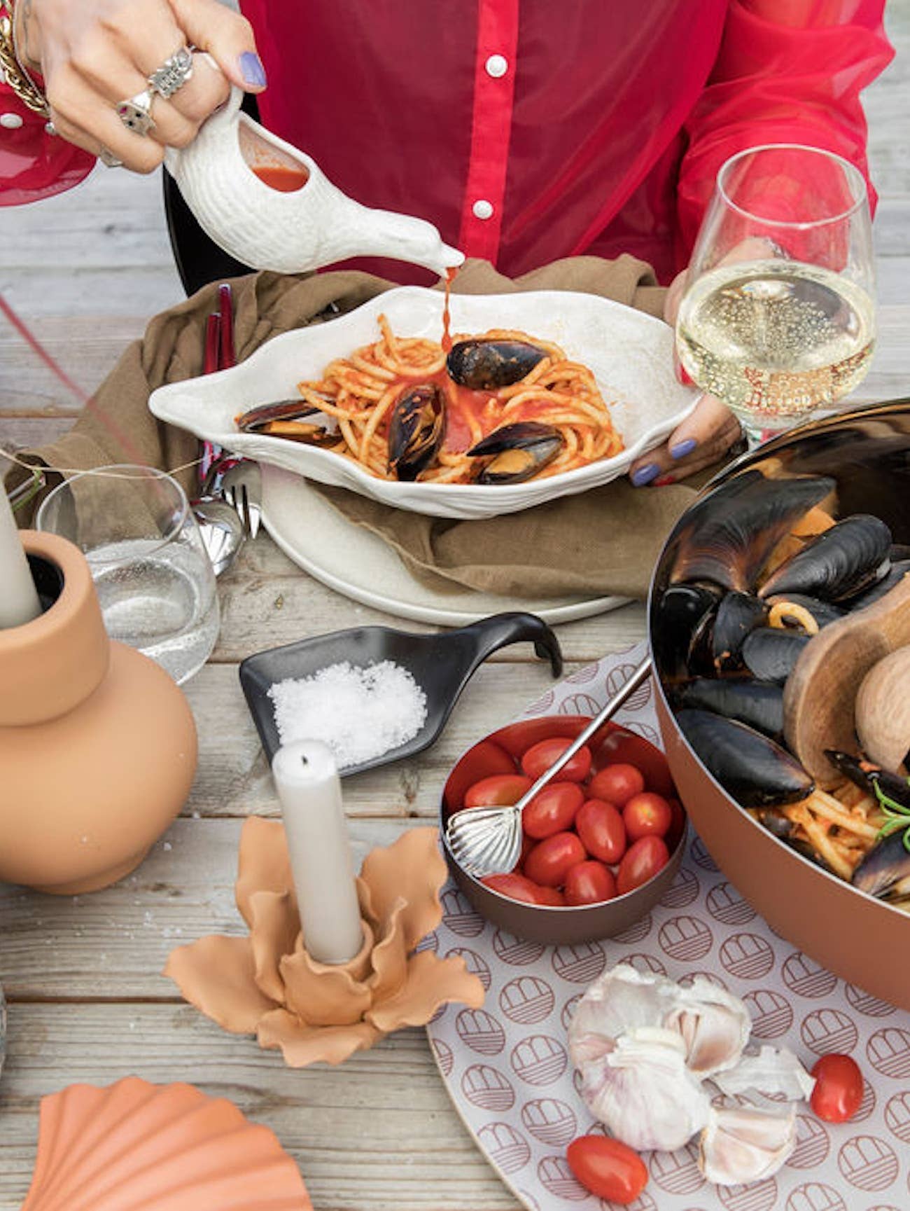 Eine Person in einem roten Hemd gießt Soße auf Spaghetti mit Muscheln, die auf der BYON | Design Schale Alima von Sagaform GmbH serviert werden, zusammen mit Weißwein, Kirschtomaten, Salz, Knoblauch und einer Kerze auf einem Holztisch.