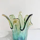Die Murano Dream | Die mittelgroße „Giusy“-Vase aus grünem Murano-Glas (H 24, Ø 18 cm) besticht durch ihren gewellten, blütenartigen Rand in Grün- und Blautönen und wird auf weiß marmoriertem Untergrund vor hellem Hintergrund präsentiert.