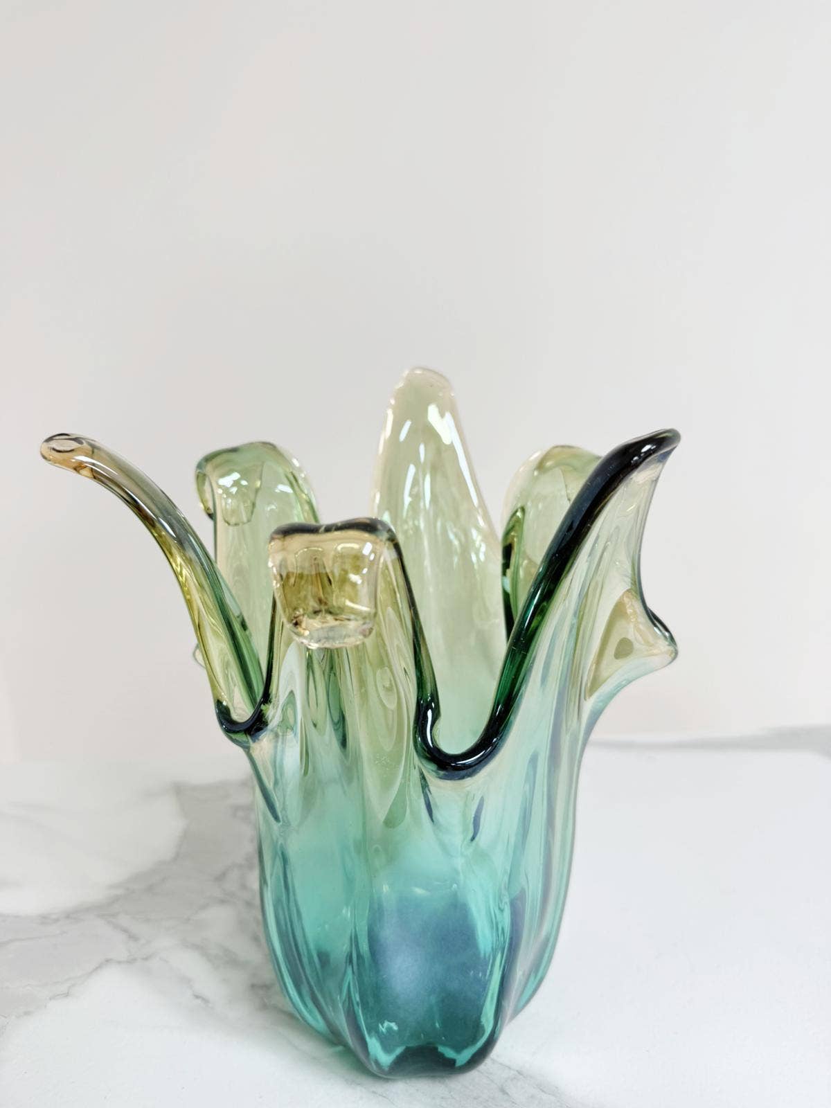 Die Murano Dream | Die mittelgroße „Giusy“-Vase aus grünem Murano-Glas (H 24, Ø 18 cm) besticht durch ihren gewellten, blütenartigen Rand in Grün- und Blautönen und wird auf weiß marmoriertem Untergrund vor hellem Hintergrund präsentiert.