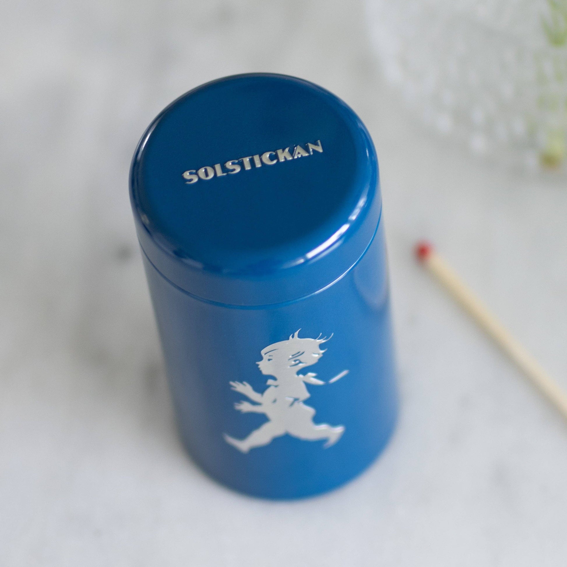 Eine Solstickan Design | Streichholzdose Blau, eine blaue zylindrische Streichholzschachtel von Solstickan Design mit der weißen Silhouette eines laufenden Kindes und SOLSTICKAN auf dem Deckel, wird auf einer hellen Fläche neben einem Holzstreichholz gezeigt.