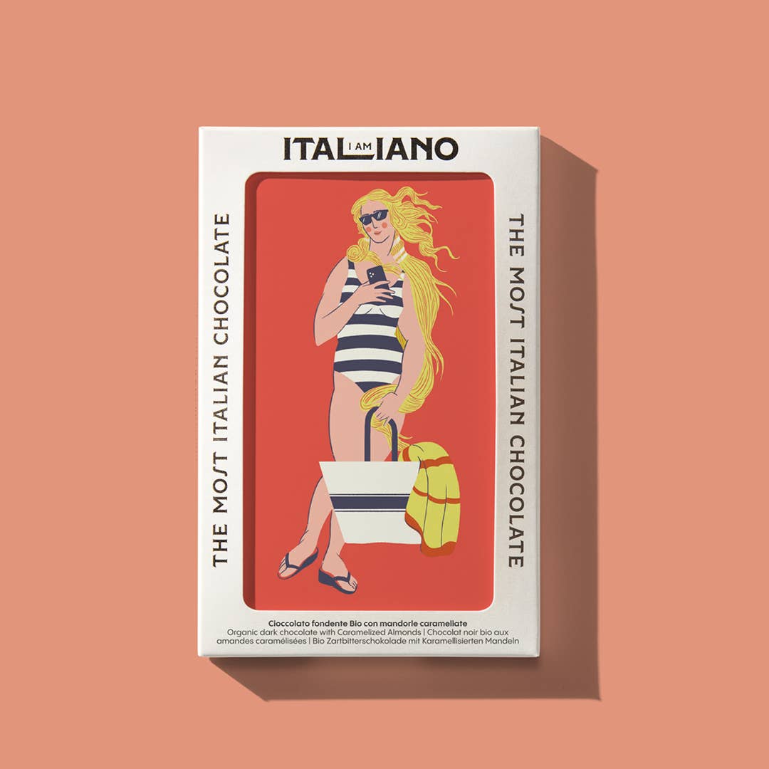 Die Schachtel I AM ITALIANO 'Venere' Bio Zartbitterschokolade mit Mandel zeigt eine stilvolle Frau mit Gelato und einer gestreiften Tasche sowie "ITALIANO" und "The Most Italian Chocolate" auf Pfirsich, wobei karamellisierte Mandeln in Bio-Zartbitterschokolade hervorgehoben werden.