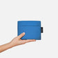 Eine Hand hält die „Notabag | ‚Original Blue‘“-Tasche von Notabag – eine kompakte, rechteckige blaue Tragetasche, die gleichzeitig als Rucksack und Handtasche dient – mit einem Deckel und einem kleinen schwarzen Etikett auf der rechten Seite vor einem schlichten weißen Hintergrund.