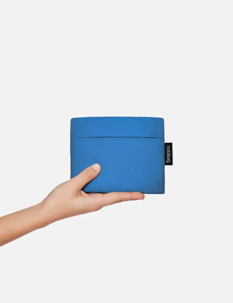 Eine Hand hält die „Notabag | ‚Original Blue‘“-Tasche von Notabag – eine kompakte, rechteckige blaue Tragetasche, die gleichzeitig als Rucksack und Handtasche dient – mit einem Deckel und einem kleinen schwarzen Etikett auf der rechten Seite vor einem schlichten weißen Hintergrund.