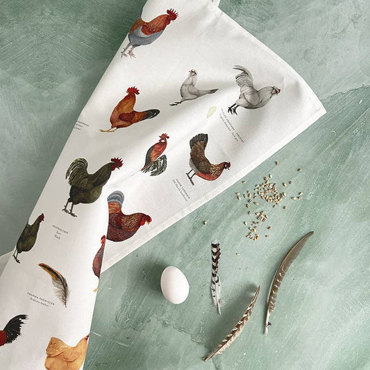 Ein Küchentuch von Koustrupco aus 100 % Bio-Baumwolle mit Hühnermotiven liegt auf einer grünen Fläche neben einem Ei, Hühnerfutter und drei Federn – ein nachhaltiges Küchenaccessoire von Koustrupco, das Stil und Funktionalität vereint.