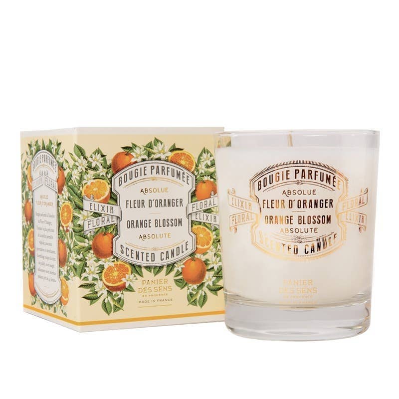 Die Glaskerze Panier Des Sens Duftkerze | Orangenblüte 180 g mit einem blumigen, von Grasse inspirierten Duft sitzt neben ihrer mit Orangenblüten- und Fruchtillustrationen geschmückten Schachtel. Diese handgefertigte vegane Duftkerze ist französischen Ursprungs.