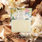 Der Panier Des Sens | Duftseife Musc des Sables 150 g ruht auf cremigen Blütenblättern mit seiner dekorativen Schachtel dahinter. Fleurs Blanches, Santal, Musc Blanc. Perfekt als festliche Badeseife für Ihr Ritual.