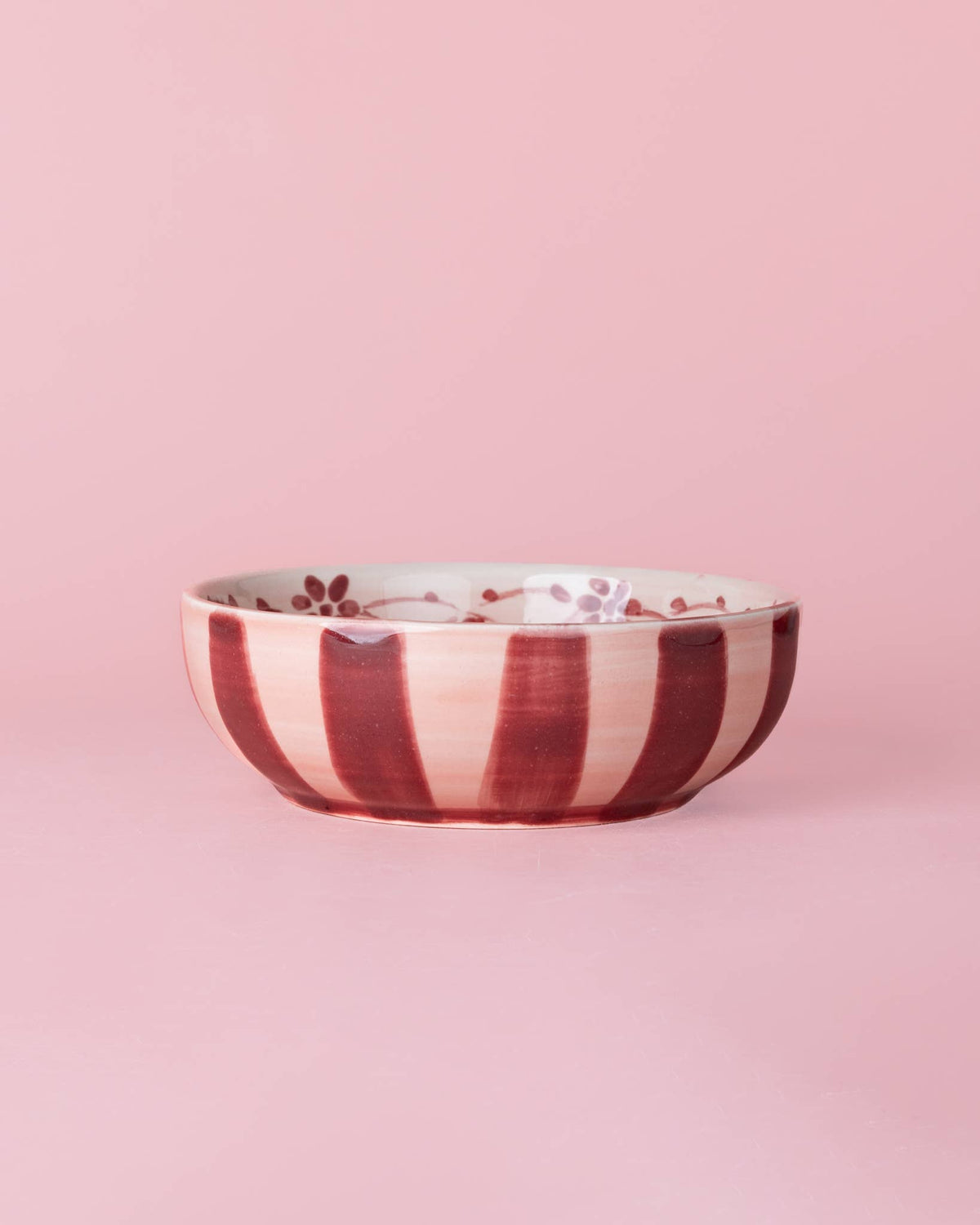 Die Alfar Tierra Cocida | Keramikschale mit Knoblauchreibe in Mittelrosa „Sakura“ von Alfar Tierra Cocida, handbemalt in Spanien, besticht durch vertikale rote und weiße Streifen sowie feine rote Blütendetails auf der Innenseite. Sie steht auf einer rosa Oberfläche vor einem rosa Hintergrund und verleiht jeder Sammlung von Küchenutensilien einen Hauch von Kunstfertigkeit.