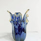 Die Vase „Giusy“ der Serie „Murano Dream“ aus blauem Murano-Glas (H 24 Ø 19 cm) zeichnet sich durch wellenförmige, ausladende Ränder und eine glatte, spiegelnde Oberfläche aus. Sie präsentiert italienische Glaskunst auf einer weißen Marmorfläche vor einem schlichten weißen Hintergrund.