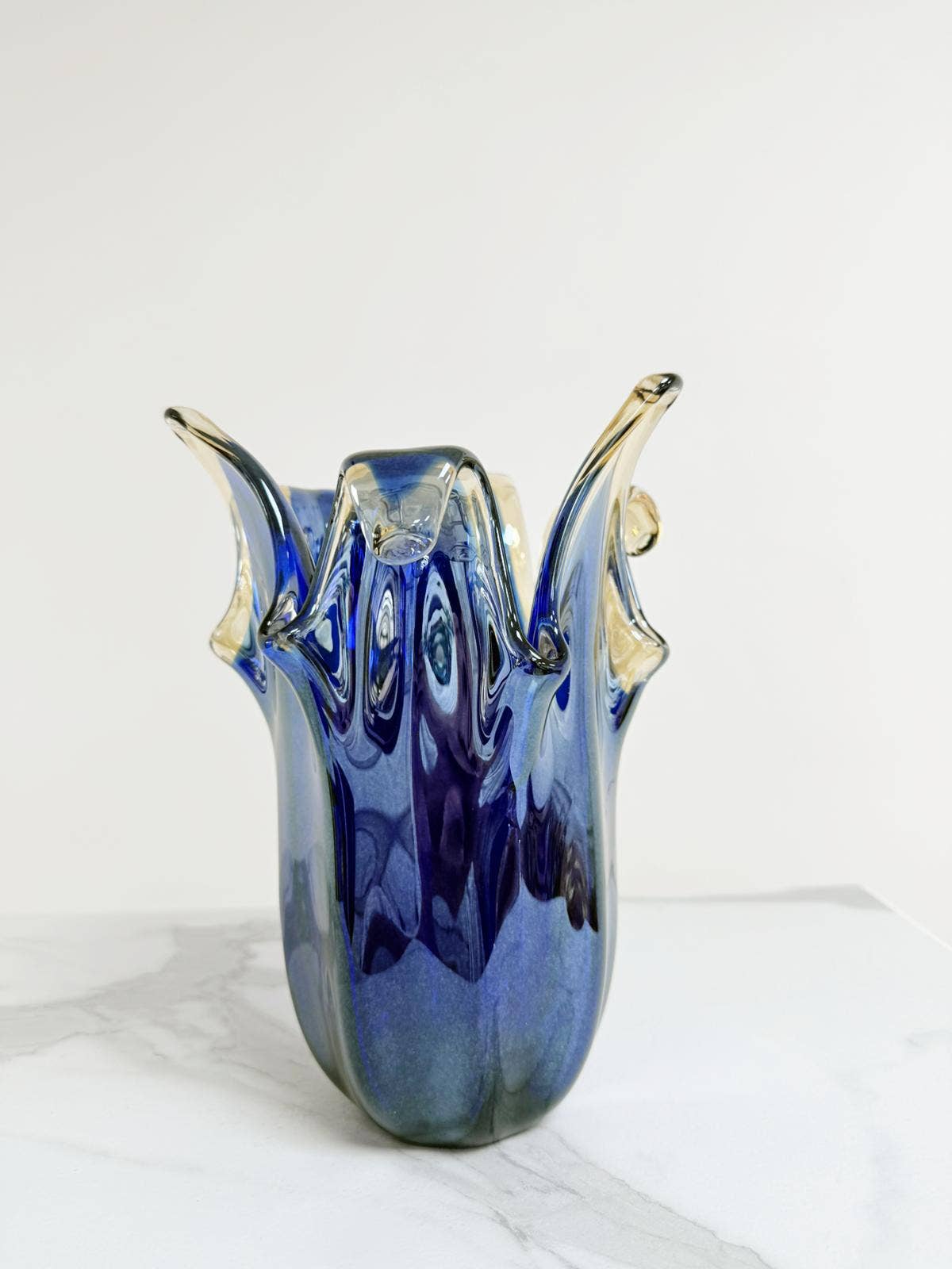 Die Vase „Giusy“ der Serie „Murano Dream“ aus blauem Murano-Glas (H 24 Ø 19 cm) zeichnet sich durch wellenförmige, ausladende Ränder und eine glatte, spiegelnde Oberfläche aus. Sie präsentiert italienische Glaskunst auf einer weißen Marmorfläche vor einem schlichten weißen Hintergrund.