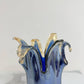 Eine mittelgroße Vase „Giusy“ aus blauem Murano-Glas (H 24, Ø 19 cm) mit gewelltem, ausladendem Rand steht auf weißem Marmor vor grauem Hintergrund. Ihre Form erinnert an fließendes, geschmolzenes Glas.