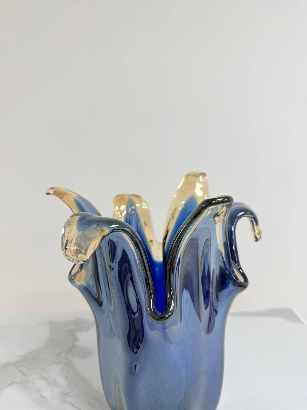 Eine mittelgroße Vase „Giusy“ aus blauem Murano-Glas (H 24, Ø 19 cm) mit gewelltem, ausladendem Rand steht auf weißem Marmor vor grauem Hintergrund. Ihre Form erinnert an fließendes, geschmolzenes Glas.