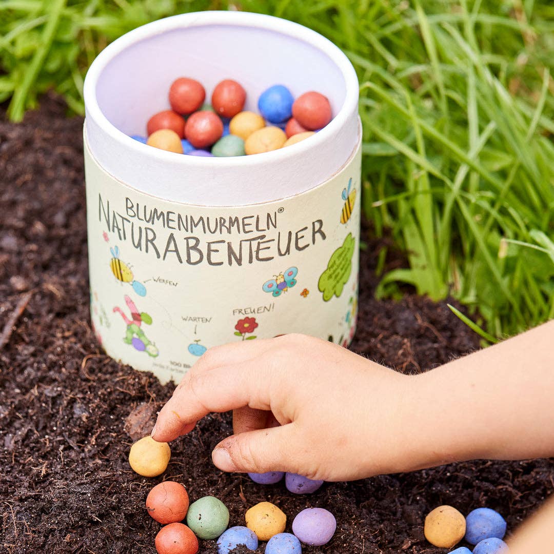 Eine Kinderhand hebt einen bunten Samenball aus der Erde neben einem runden Behälter mit der Aufschrift „Stadtgärtner Saatgut | Blumenmurmeln ‚Naturabenteuer‘ 100er-Dose“ auf, der mit weiteren bunten Samenbällen gefüllt ist und von grünem Gras umgeben ist– ein ideales nachhaltiges Geschenk für Kinder und ein spannendes Naturabenteuer für kleine Entdecker.