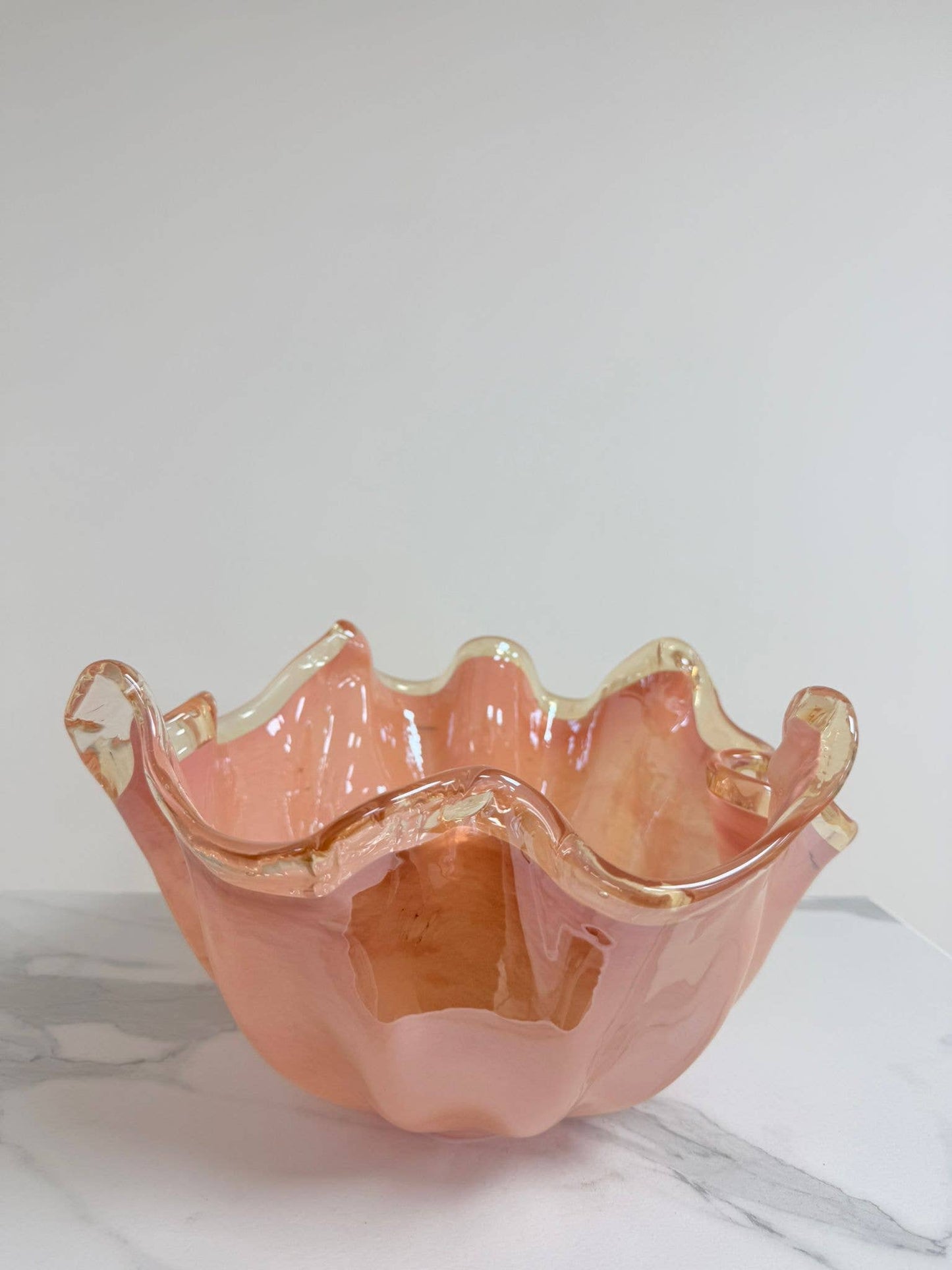 Die Murano Dream Dekoschale „Fazzoletto“ aus rosa Murano-Glas (H 20 cm, Ø 22 cm) mit gewellten Rändern und goldenem Rand glänzt auf einer weißen Marmorfläche vor einem schlichten, hellen Hintergrund.