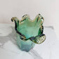 Die mittelgroße Vase „Venezia“ aus grünem Murano-Glas (H 25, Ø 14 cm) von Murano Dream mit gewellten Kanten und klaren Akzenten steht auf weißem Marmor vor hellem Hintergrund.