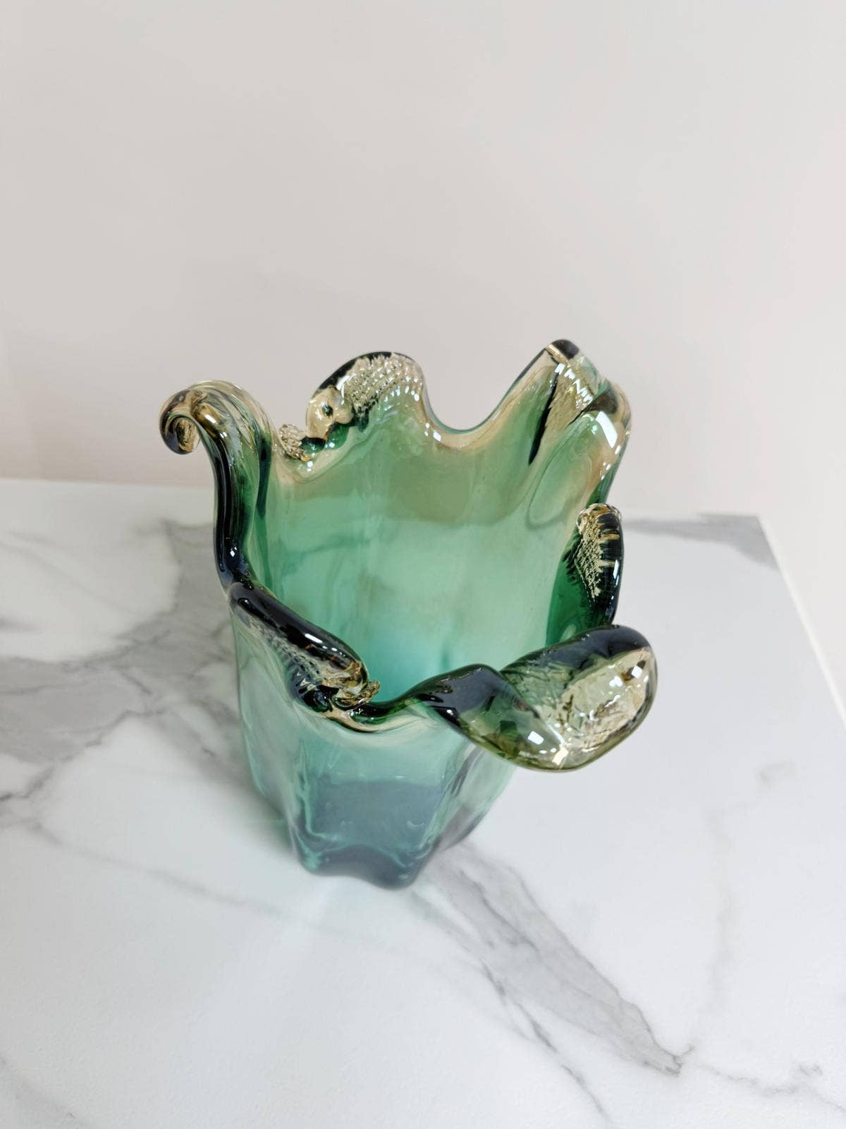Die mittelgroße Vase „Venezia“ aus grünem Murano-Glas (H 25, Ø 14 cm) von Murano Dream mit gewellten Kanten und klaren Akzenten steht auf weißem Marmor vor hellem Hintergrund.