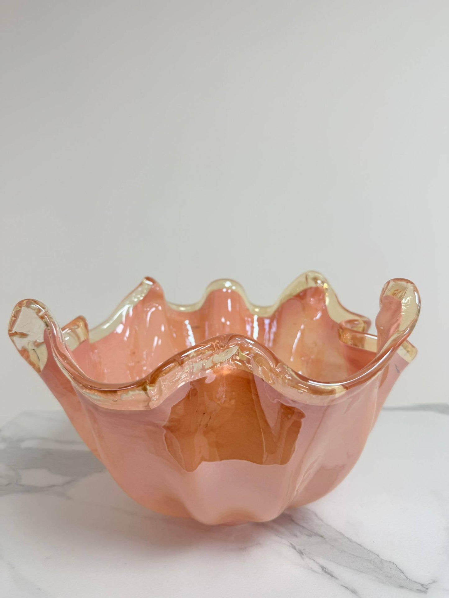 Die Murano Dream Dekoschale „Fazzoletto“ aus Murano-Glas in Rosa (H 20, Ø 22 cm) besticht durch gewellte, durchscheinende Ränder und präsentiert feinste italienische Glaskunst auf einer weiß-grau marmorierten Oberfläche vor hellem Hintergrund.