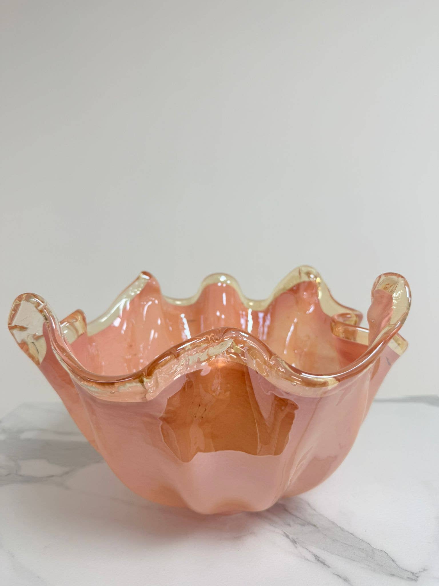 Die Murano Dream Dekoschale „Fazzoletto“ aus Murano-Glas in Rosa (H 20, Ø 22 cm) besticht durch gewellte, durchscheinende Ränder und präsentiert feinste italienische Glaskunst auf einer weiß-grau marmorierten Oberfläche vor hellem Hintergrund.