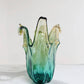 Die Murano Dream „Giusy“-Vase (H 24, Ø 18 cm) besticht als handgefertigte, mittelgroße Vase aus grünem Murano-Glas mit eleganten Farbverläufen in Grün, Gelb und Klarglas sowie durch ihre abgerundete, organische Form.