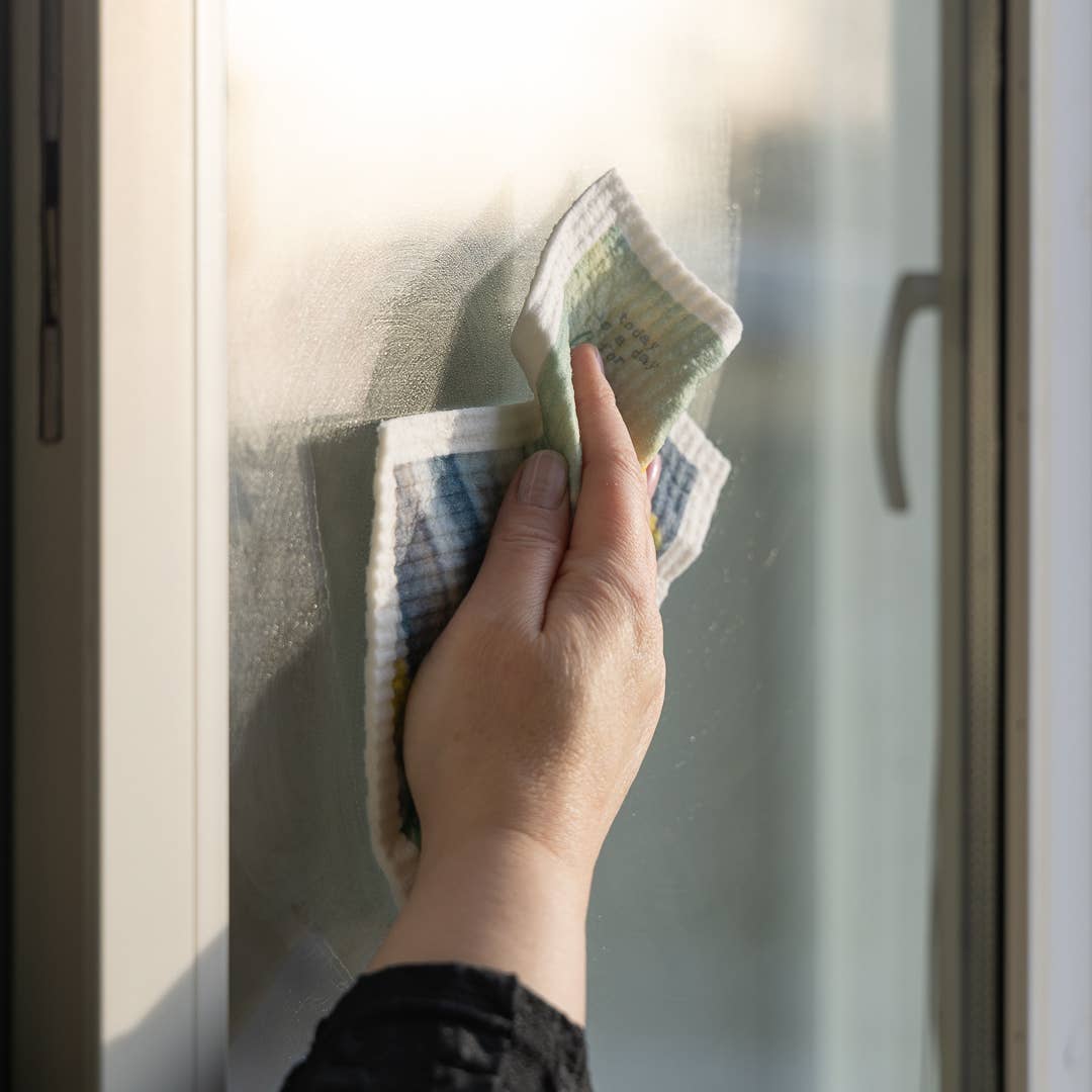 Eine Hand wischt ein Glasfenster mit dem More Joy Home | 'Rote Tulpen' Biologisch abbaubares Schwammtuch. Sonnenlicht strömt herein und erzeugt eine sanfte Reflexion, während dieses Zero-Waste-Produkt von More Joy Home die Oberfläche reinigt.