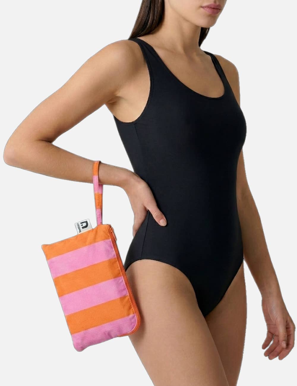 Eine Frau in einem schwarzen, ärmellosen Badeanzug hält die „Notabag | Handtuch orange & rosa gestreift 78 × 160 cm“ in der Hand, ein leichtes Reisehandtuch aus recyceltem Polyester mit Reißverschluss und Trageschlaufe. Der Hintergrund ist schlicht hellgrau.