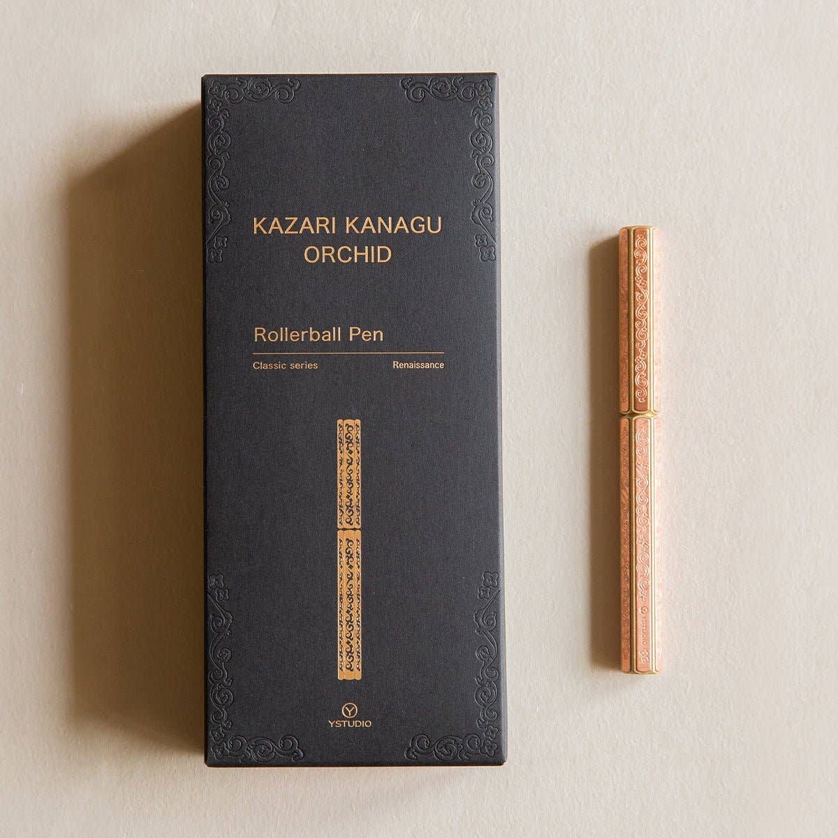 Ein schwarzer YSTUDIO | Klassische Renaissance KAZARI KANAGU ORCHID Tintenroller von ENTREPÔT ruht neben einem goldenen, kunstvoll gemusterten Stift mit exquisiter Metallgravurkunst auf einer beigen Oberfläche.