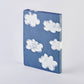 Das nuuna „Head In The Clouds“ Notizbuch Graphic L A5+ von nuuna steht aufrecht in einem blauen Hardcover-Einband und ist mit verspielten weißen Comic-Wolken verziert, die auf der Vorderseite und dem Buchrücken vor einem schlichten, hellen Hintergrund verteilt sind. Dieses Notizbuch aus recyceltem Denim und hochwertigem Papier verbindet umweltbewusste Materialien mit einem verspielten Design.