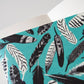 Aus dem Papaya Paper Products | Notizheft A5 mit Federmuster blau von Papaya Paper Products lugt ein tealfarbener Umschlag mit schwarz-weißem Federmuster hervor.