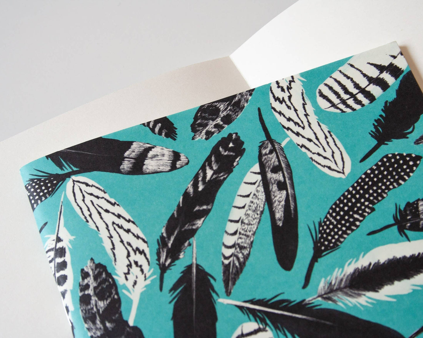 Aus dem Papaya Paper Products | Notizheft A5 mit Federmuster blau von Papaya Paper Products lugt ein tealfarbener Umschlag mit schwarz-weißem Federmuster hervor.