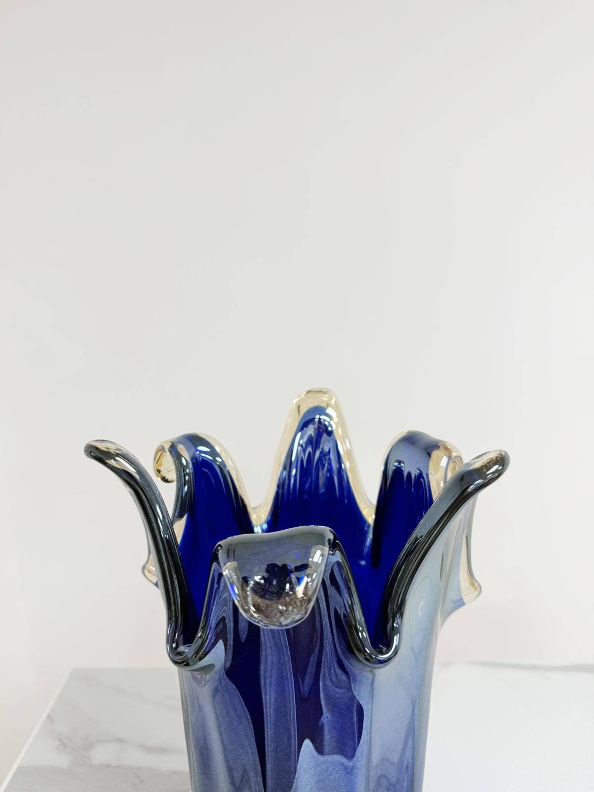 Die Murano | Große Giusy-Vase aus blauem, originalem Murano-Glas von Murano Dream mit welligen, geschwungenen Rändern steht auf weißem Marmor vor einem schlichten Hintergrund. H 32 cm, Ø 20 cm.