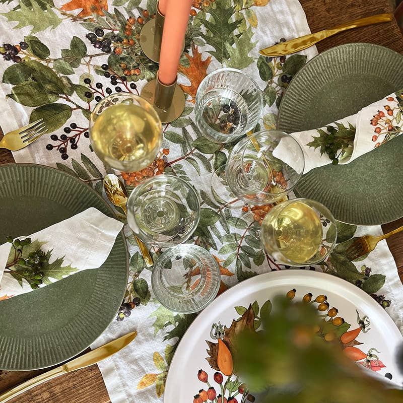 Ein für zwei Personen gedeckter Tisch mit grünen Tellern, goldenem Besteck, Weingläsern sowie einer Tischdecke und Servietten mit herbstlichem Blumenmuster wird durch den Koustrupco | Tischläufer aus Leinen 145 x 45 cm im herbstlichen Design wunderschön ergänzt, der der herbstlichen Tischdekoration eine elegante Note verleiht. In der Mitte steht eine rosa Kerze in einem goldenen Kerzenhalter.