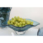 Aulica | Dekorative blaue Glasschale 33 x 18 x 10,5 cm von AULICA, gefüllt mit grünen Trauben, steht auf einer weißen Oberfläche. Im Hintergrund ist eine weitere Schale teilweise sichtbar.