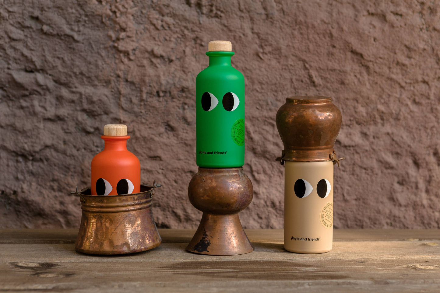 Drei leuchtende Flaschen mit verspielten Augen - orange, grün und beige - ruhen auf kupfernen Gefäßen vor einer braunen Wand und präsentieren das Natives Olivenöl extra mit Basilikum 200 ml von yiayia and friends auf einer rustikalen Holzoberfläche.