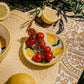 Auf dem Alfar Tierra Cocida 'Limone' Keramik Knoblauchreibe Teller (12,5 x 12,5 x 2,5 cm) liegen Kirschtomaten neben einer ganzen Zitrone, einer Zitronenhälfte und blättrigen Zweigen auf einem gemusterten Tischtuch und einem gesteppten Platzset.